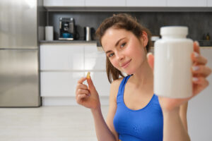 Close-up portret van jonge vrouw, fitnessinstructeur die een fles vitamines laat zien, die een dieetpil neemt, thuis zit en oefeningen doet
