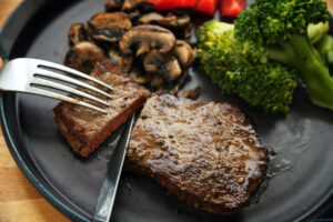 biefstuk wordt met mes en vork gesneden, met groenten zoals broccoli, champignons en tomaten, koolhydraatarm dieetdiner op een donkergrijs bord, geselecteerde focus, geringe scherptediepte
